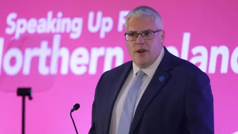 Gavin Robinson - A man with grey hair wearing glasses. Dressed in a navy blue suit and light blue tie, with a blue flower pin on the right hand side of blazer.