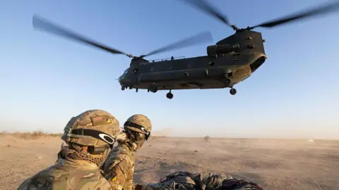 Two joint helicopter support squadron soldiers look away from camera while wearing camo gear and helmets, with a CH-47 Chinook mid air above them in a desert type environment. Image taken near El Centro USA in October.