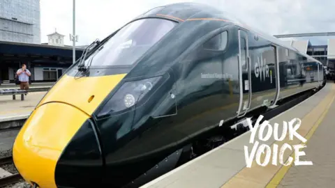 A GWR train is seen at at station with a man with a camera standing on a platform behind. Your Voice branding is in the corner of the photograph. 