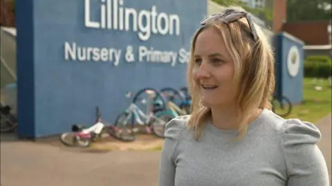 A woman outside a primary school, with bikes propped against a wall, is wearing a grey blouse. She has sunglasses resting on the top of her head and is smiling as she talks to a reporter out of view on the left hand side of the image.