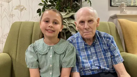 An eight-year-old girl in green school uniform sits next to an 86-year-old man. The girl has brown hair pulled back in a ponytail while Dennis wears a blue and white checked shirt and dark trousers. They sit on a green sofa.