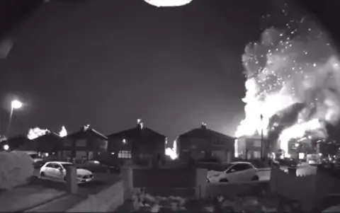 A black and white screenshot shows huge flames coming out of a home on the right hand side of the image. There is a row of home on the road opposite the camera