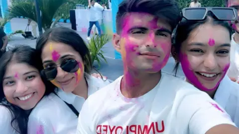 TheBiGhitentertainment Three women and a man wearing white tops are covered with bright pink powder as part of the Holi celebrations. They are all smiling.