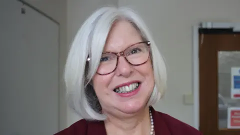 A woman with white hair and tortoiseshell glasses smiles at the camera, she is wearing pears and a maroon blazer. Behind her is a brown office door.