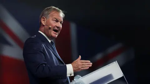 Reuters Rupert Lowe is standing at a podium giving a speech. He's wearing a dark blue jacket, white shirt and blue striped tie. Behind him, we can see a Union Flag.