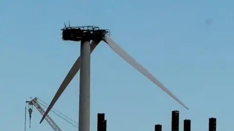 Andrew Turner/BBC A burned shell of a turbine housing on an offshore wind farm. The uppermost blade has been removed, with two in the downward position. A crane jib and struts from a jack-up barge are visible in the lower part of the image. The sky behind the semi-silhouetted image is blue.