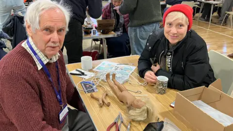 A man and a woman are sat across from one another at a table.  Both are looking to camera.  The man is on the left and is wearing a dark red jumper over a white shirt.  He has a lanyard around his neck.  The woman is on the right and wears a black coat, and a red wooly hat.  On the table between them is a plastic doll with blonde hair. The doll is missing its arms and is in the process of being fixed.  Also on the table are the arms of the doll, bits of string and pairs of pliers needed for the repair.