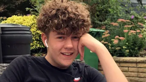 Stephens Family Olly Stephens has curly dark hair and is wearing a black t-shirt. He is sat down smiling.