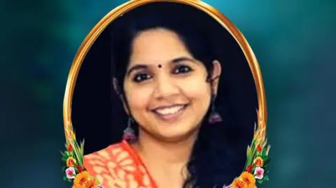 Union of UK Malayalee Associations Ms Renjitha smiles at the camera, wearing traditional costume. The photo is contained in an oval shape, bordered by painted flowers and a gold-coloured frame.