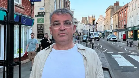 Mr Chitty, who wears a white t-shirt, cream shirt and clear glasses, looks at the camera. The photo has been taken in central Well, with the High Street and cathedral visible behind him.