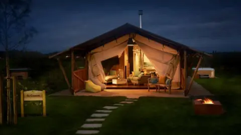 Love2Stay A small wooden lodge in the dark, with the front lit up and some furniture seen at the front, with a stone path in the foreground