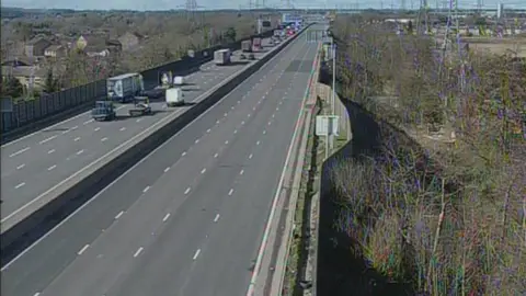 Two carriageways of a motorway. Cars and lorries can be seen in the distance on the left side, while the right side is empty. There are houses and trees either side of the motorway that is slightly elevated. 