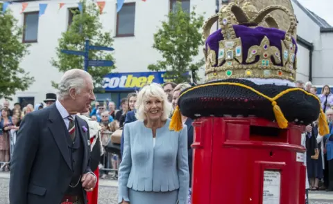 PA Media King Charles and Queen Camilla admire a post box decorated with a crocheted crown
