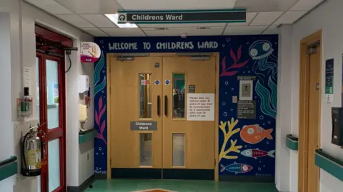 Double doors at the end of a corridor with "Welcome to the Children's Ward" written above them with an under the sea mural on the surrounding wall.