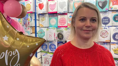 A woman with a blonde bob and a red knitted jumper smiles as she stands in a balloon shop. There is a gold star-shaped balloon with writing on beside her and behind her is a wall full of packets of balloons.