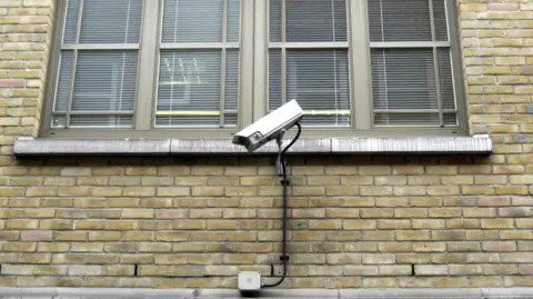 The side of a brick building. There is a white CCTV camera attached to it in front of a large window.
