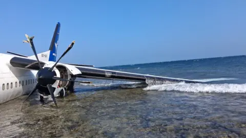 Starsky Airlines A damaged aircraft rests partly submerged on a shallow shoreline, with waves washing against its wing as it lies in clear, shallow water under a bright blue sky.