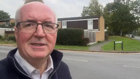 A man with balding grey hair and thin black-framed glasses is standing in a residential street, wearing a blue jumper and a white shirt.