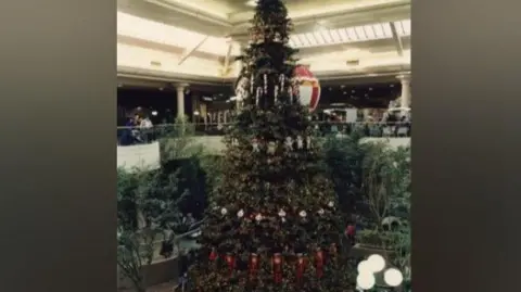 Metrocentre A giant Christmas tree in the centre of a large atrium. It is decorated with red and white decoations including candy canes, nutcrackers and fairy lights. There are other trees around it and people milling around at the bottom. There is a balcony above the main floor and the ceiling is covered in glass.
