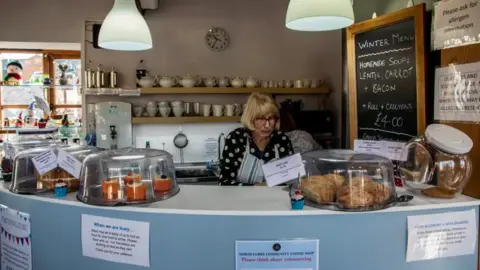 North Curry Coffee Shop A volunteer behind the counter with piles of fresh cakes in the foreground.