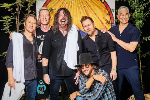 Tom Jackson The Foo Fighters standing together looking at the camera smiling. They are all sweaty and two have white towels on their shoulders including Dave Grohl. He has his mouth open is a wide smile and his long curly hair is down in front of his left eye