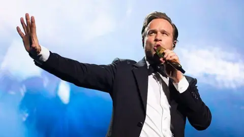 Getty Images Olly Murs dressed in a black suit and white shirt sings into a mic during one of his concerts