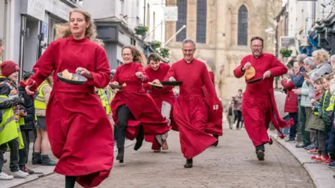 PA Media Two women and three men in red cassocks run down a street holding frying pans with crepes in them. 