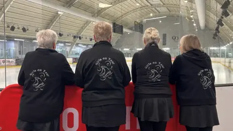 Four of the skaters have their backs to the camera looking at the ice rink. They are wearing black jackets with their Girls on Tour logo embroidered on the back in white. 