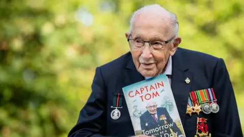 Getty Images Captain Sir Tom Moore wearing his military medals holding a copy of his autobiography. He has receding white hair, a navy blazer, and there are trees in the background