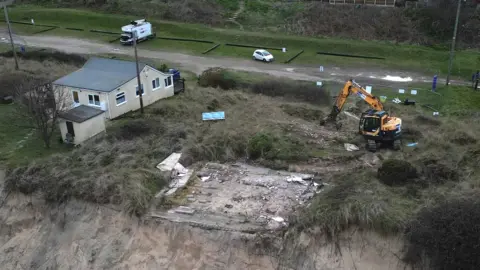 The site of a demolished house, next to a JCB and another home that is still standing