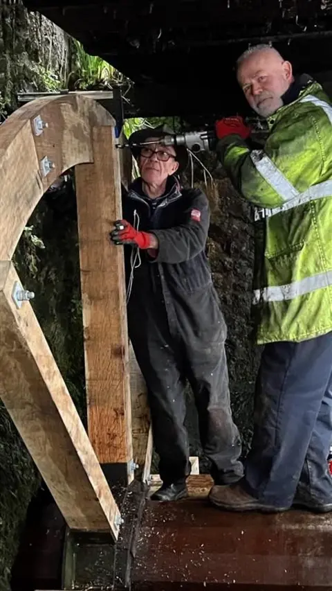 A man wearing a high-vis jacket and drilling into a wooden waterwheel. Another person is holding the waterwheel still.