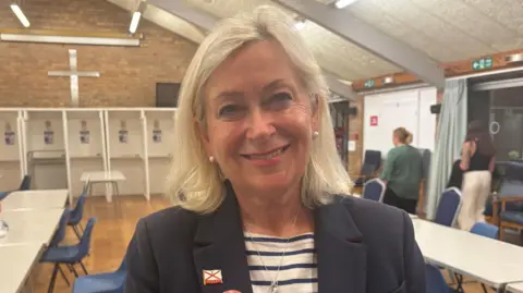 Helen Miles smiles at the camera from a hall where election votes have been counted in Jersey. She has shoulder-length white hair with a tinge of blonde. She has silver ear-rings on and a silver necklace. She is wearing a dark blazer with a navy blue and white striped shirt underneath. A Jersey flag badge is on her right lapel.