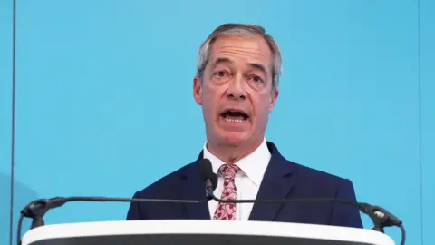 PA Media Reform UK leader Nigel Farage stands at a lectern delivering a speech. He wears a white shirt with a bright pink and red tie and a navy suit jacket. He has grey cropped hair.