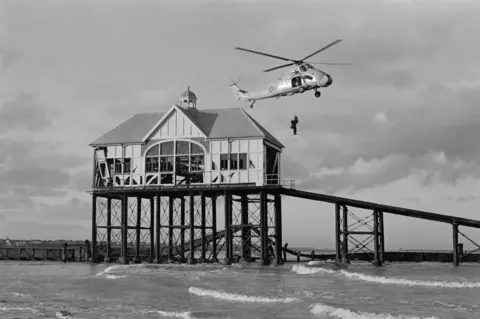 A black and white photograph shows an RAF rescue helicopter hovering above the gentle waves of the sea. A person hangs below the helicopter on a winch. In the background, the damaged remains of the RNLI boathouse can be seen.
