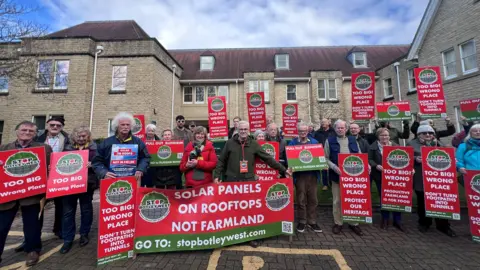 Protesters from the Stop Botley West group outside West Oxfordshire District Council's headquarters holding signs, most of which say: "Too big! Wrong place. Protect our heritage." 