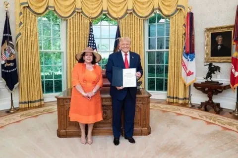 Dame Karen Pierce Dame Karen Pierce standing in the Oval Office with Donald Trump. She is wearing an orange outfit and a hat. Trump is wearing a suit and red tie and holding a folder towards the camera, as he smiles.