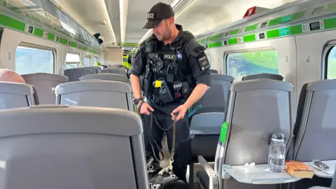 BBC A police officer and a black sniffer dog on a train checking passengers fro drugs