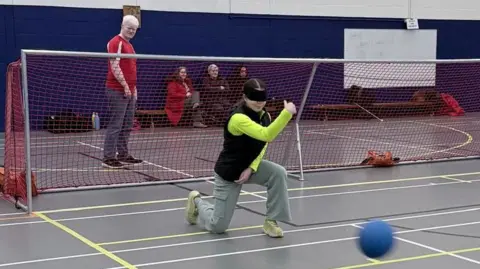 Goalball players are taking part in a taster session. They are wearing eyeshades. One player is on one knee after throwing the ball. Another player is sitting on the floor. Spectators are nearby. There is a net across the court.