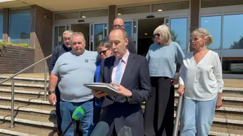 Charlie Rose/ BBC A man, Paul Stanley, stood on steps outside a court building reading a statement on a piece of paper. A group of six people are stood in a line behind him.