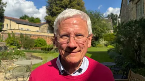 BBC Colin Fletcher smiles for a photo taken outside his house. He has neat white hair and glasses. He is wearing a white collared shirt with a red round-necked pullover on the top. His lawn and garden furniture can be seen in the background.