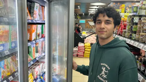 A man wearing a hoodie pictured next to a fridge in a supermarket.