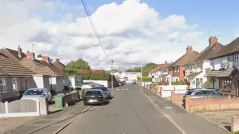 Google Houses are on either side of the road with cars parked outside a few of them and on the pavement. The sun is shining.