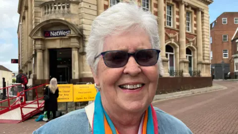 Headshot of a woman with white hair, smiling, wearing dark sunglasses and a colourful scarf standing outside a high street bank