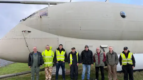A group of men standing in front of a vintage plane on an airfield