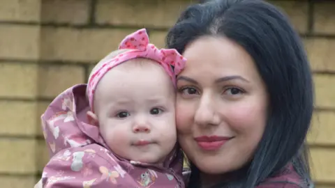NHS Lanarkshire Dr Leila El Alti holds her baby Aurora up while standing outside. Dr El Alti has dark hair to her shoulders, while her baby is wearing a violet top and a pink bow on her head. 