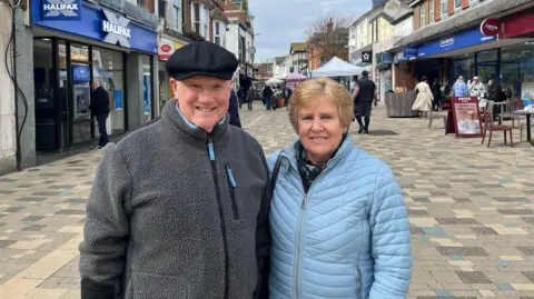 George Carden/BBC A man in a grey fleece wearing and a black hat, and a woman wearing a light blue puffer jacket.