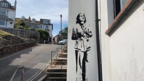 A mural of Barbara Hepworth painted on to a plain white wall. It's on a narrow and steep street in St. Ives with steps on one side.