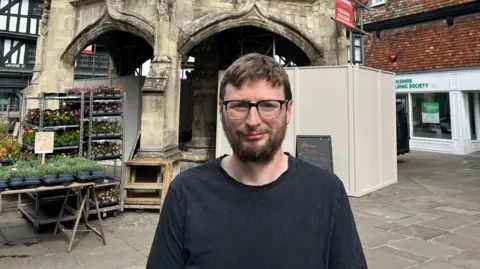 Councillor Sam Charleston looks at the camera with the Poultry Cross in the background. He is wearing glasses, has a dark long-sleeved top on and has a dark beard and short dark hair