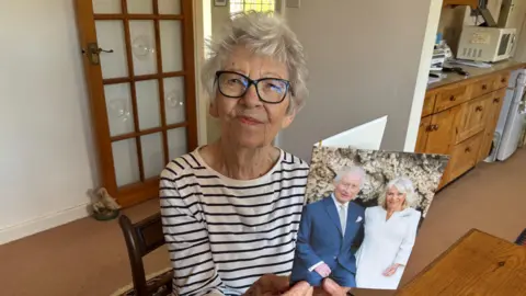 Luke Deal/BBC An elderly woman sits at a kitchen table. She is looking at the camera and smiling. She holds a card with the images of King Charles III and Queen Camilla on the front. 