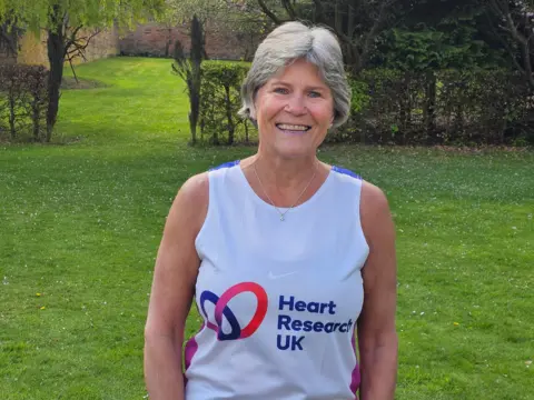 Janine Anderson A woman is stood in a garden, visible from the waste up. She has a short grey bob and is smiling at the camera. Her top is a white vest and reads "Heart Research UK".